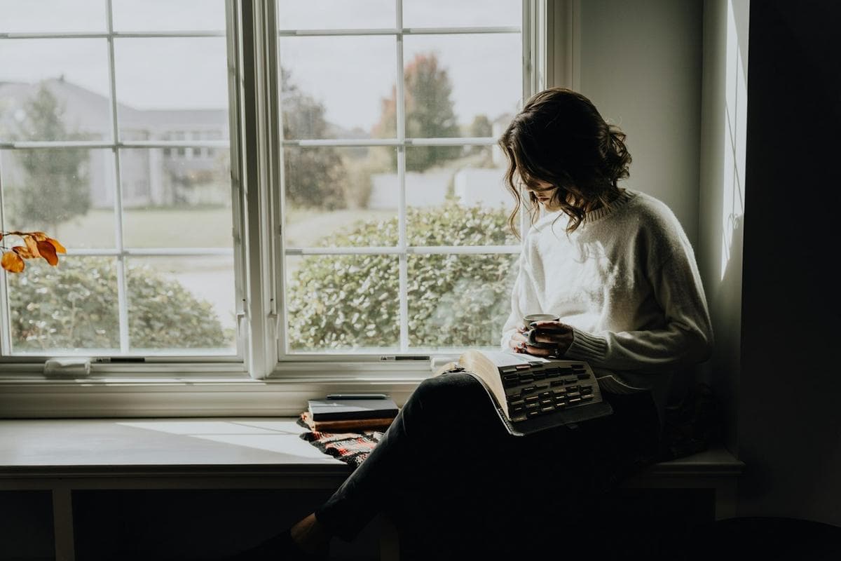A woman reading in a sunny window seat with a mug.